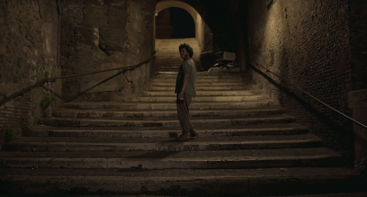 A young man stands on a long stone stairway beneath an archway at night