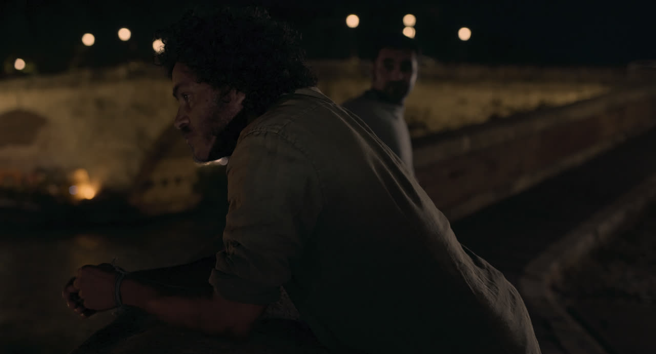A young man sits on a riverside wall under Rome’s night lights